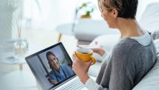 Person sitting on a couch speaking to a healthcare professional on a computer while holding a cup in their hand