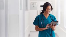 Nurse walking down a white hallway while wearing scrubs and looking at a tablet