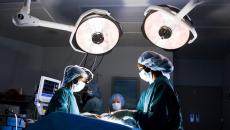 Healthcare providers in an operating room looking down at a patient on a table with large lights above them 