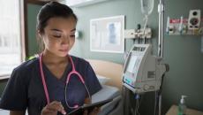 Healthcare provider standing in a hospital room wearing a pink stethoscope while holding a tablet 